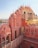 An iconic palace featuring intricate pink sandstone architecture with multiple arched windows and domes. Several people are observing the view from a balcony. The structure showcases traditional craftsmanship with detailed patterns and embellishments.