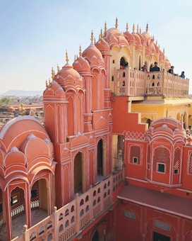 An iconic palace featuring intricate pink sandstone architecture with multiple arched windows and domes. Several people are observing the view from a balcony. The structure showcases traditional craftsmanship with detailed patterns and embellishments.