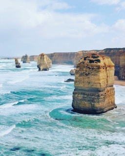 Large limestone rock formations stand majestically in the ocean, surrounded by turquoise waves crashing against them. The coastline features steep cliffs with layers of rock visible. The sky is partly cloudy, giving a bright but slightly overcast atmosphere.