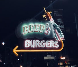 A brightly lit neon sign for 'Beep's Burgers' with a rocket design, glowing against a dark night backdrop. A yellow arrow points to the left beneath the text.