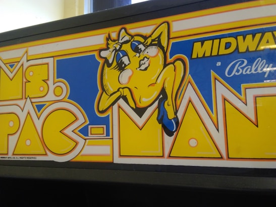 A colorful arcade game marquee displaying a stylized character resembling a yellow circle with eyes, arms, and legs. The character is wearing blue shoes and appears playful with expressive facial features. Bright colors and bold lettering highlight the game title 'Ms. Pac-Man' along with branding from Midway and Bally.