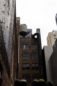 An urban scene featuring a tall brick building with multiple windows. A cylindrical water tower is perched on the rooftop along with some metallic structures and pipes. Adjacent buildings, mostly modern and made from metal or concrete, are visible in the background. Below, there are flowers in planters adding a touch of greenery.
