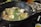 A large black pot filled with a variety of ingredients simmering in broth. Visible ingredients include broccoli, green beans, mushrooms, tofu, and assorted vegetables. The setup suggests a communal dining experience, possibly a hot pot meal.