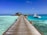A wooden boardwalk extends over clear turquoise waters, leading to a thatched-roof structure over the ocean. Large potted plants line the pathway, and a white boat labeled 'parasailing' is moored in the water nearby.