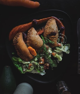 A plate with three empanadas arranged artistically on a base of red sauce. Fresh lettuce, diced tomatoes, and herbs surround the empanadas, adding a touch of green. A carrot slice and some dill garnish the dish. Nearby, an avocado and a carrot are placed on the dark surface, adding a rustic ambiance.