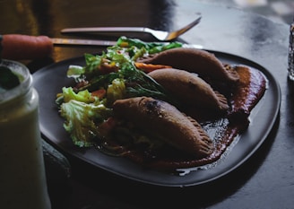A plate with empanadas and a fresh salad, garnished with a rich sauce, is placed on a dark table. Nearby, there is a smoothie in a glass jar and various utensils.