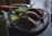 A plate with empanadas and a fresh salad, garnished with a rich sauce, is placed on a dark table. Nearby, there is a smoothie in a glass jar and various utensils.