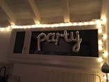 A string of fairy lights is arranged around a window, illuminating the word 'party' formed with metallic balloons. The setting appears to be indoors with white walls and a ceiling with rafters.