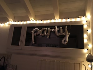 A string of fairy lights is arranged around a window, illuminating the word 'party' formed with metallic balloons. The setting appears to be indoors with white walls and a ceiling with rafters.