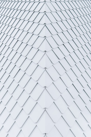 Uniformly arranged white tiles create a geometric pattern with a symmetrical and seamless appearance, resembling a roof or wall surface. The tiles are aligned in diagonal rows, each slightly overlapping the next, forming a repetitive and organized texture.
