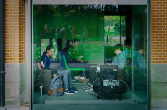 A group of people are seated inside a glass-walled room, some using laptops and mobile devices. The interior and glass reflect green hues from the surroundings, creating an almost surreal atmosphere. The setting is calm, suggesting a study or work environment, possibly within an urban park or garden due to visible greenery outside.