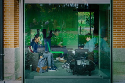 A group of people are seated inside a glass-walled room, some using laptops and mobile devices. The interior and glass reflect green hues from the surroundings, creating an almost surreal atmosphere. The setting is calm, suggesting a study or work environment, possibly within an urban park or garden due to visible greenery outside.