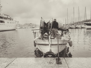 A small fishing boat docked at a marina with a person standing on the stern. The boat has various fishing equipment, and multiple other boats and yachts are anchored nearby. The background consists of a cloudy sky and distant marina infrastructure.