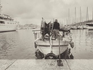 A small fishing boat docked at a marina with a person standing on the stern. The boat has various fishing equipment, and multiple other boats and yachts are anchored nearby. The background consists of a cloudy sky and distant marina infrastructure.