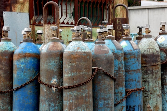 Multiple rusted industrial gas cylinders are arranged in a row, secured by chains. The tanks show significant signs of wear, with peeling paint and rust marks. They are placed in an outdoor setting, near a metal structure with a gridded window and weathered surfaces.
