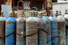 Multiple rusted industrial gas cylinders are arranged in a row, secured by chains. The tanks show significant signs of wear, with peeling paint and rust marks. They are placed in an outdoor setting, near a metal structure with a gridded window and weathered surfaces.