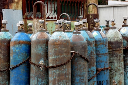 Multiple rusted industrial gas cylinders are arranged in a row, secured by chains. The tanks show significant signs of wear, with peeling paint and rust marks. They are placed in an outdoor setting, near a metal structure with a gridded window and weathered surfaces.