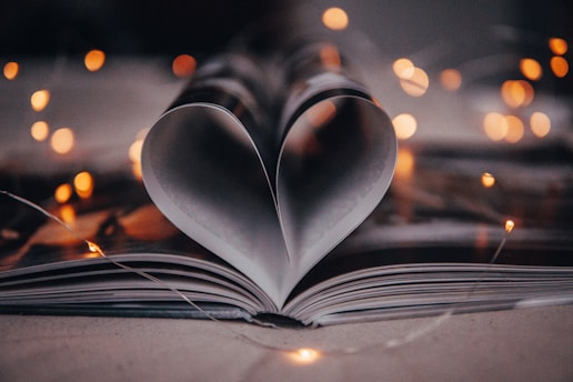 An open book has its pages creatively folded to form a heart shape in the center. Soft fairy lights create a warm bokeh effect in the background, adding a cozy and romantic ambiance.