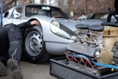 A silver vintage sports car with a person partially visible underneath the vehicle, likely performing maintenance or repair. In the foreground, there is an exposed car engine with visible mechanical components, labeled with the brand name 'Porsche.' The setting appears to be an outdoor or garage environment.