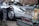 A silver vintage sports car with a person partially visible underneath the vehicle, likely performing maintenance or repair. In the foreground, there is an exposed car engine with visible mechanical components, labeled with the brand name 'Porsche.' The setting appears to be an outdoor or garage environment.