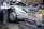 A silver vintage sports car with a person partially visible underneath the vehicle, likely performing maintenance or repair. In the foreground, there is an exposed car engine with visible mechanical components, labeled with the brand name 'Porsche.' The setting appears to be an outdoor or garage environment.