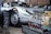 A silver vintage sports car with a person partially visible underneath the vehicle, likely performing maintenance or repair. In the foreground, there is an exposed car engine with visible mechanical components, labeled with the brand name 'Porsche.' The setting appears to be an outdoor or garage environment.