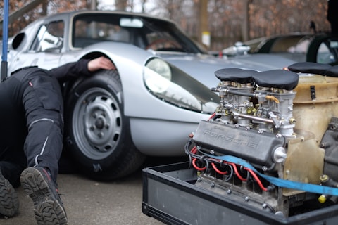 A silver vintage sports car with a person partially visible underneath the vehicle, likely performing maintenance or repair. In the foreground, there is an exposed car engine with visible mechanical components, labeled with the brand name 'Porsche.' The setting appears to be an outdoor or garage environment.