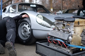 A silver vintage sports car with a person partially visible underneath the vehicle, likely performing maintenance or repair. In the foreground, there is an exposed car engine with visible mechanical components, labeled with the brand name 'Porsche.' The setting appears to be an outdoor or garage environment.
