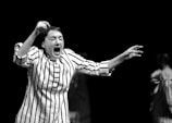 A person is expressing a strong emotion, possibly shouting or singing, with their mouth wide open. They are wearing a long-sleeved striped shirt. The background is dark, and there are blurred figures wearing similar striped garments.