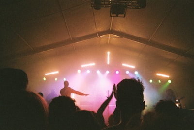 A dimly lit concert stage with colorful lights illuminating the scene. The silhouette of a performer is seen in the foreground along with the outlines of audience members. The atmosphere suggests an energetic and lively music event.