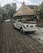 An old white convertible car is parked on a stone-paved street beside a traditional thatched roof hut. Trees and plants surround the area, giving a lush, tropical ambiance. Another white car is visible in the background parked on the same street.