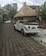 An old white convertible car is parked on a stone-paved street beside a traditional thatched roof hut. Trees and plants surround the area, giving a lush, tropical ambiance. Another white car is visible in the background parked on the same street.