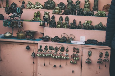A display of various metal figurines and decorative items arranged on shelves. The shelves have a pink background and hold objects like statuettes, teapots, and hanging bells. The pieces appear to be made of brass or similar metals, showcasing intricate designs and diverse forms including animals and mythical creatures.