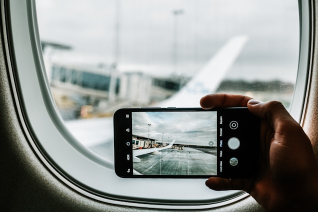 A hand holds a smartphone, capturing a view from an airplane window, showing the airport tarmac and part of the aircraft wing outside. The image on the phone screen mirrors the scene outside the window.