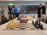 A chess tournament setting with chessboards lined up on a long table, each equipped with a set of black and white chess pieces. A digital chess timer is placed between the sets, showing a time reading. In the background, several people are standing and talking, slightly blurred due to the depth of field.
