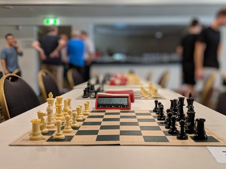 A chess tournament setting with chessboards lined up on a long table, each equipped with a set of black and white chess pieces. A digital chess timer is placed between the sets, showing a time reading. In the background, several people are standing and talking, slightly blurred due to the depth of field.