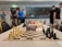 A chess tournament setting with chessboards lined up on a long table, each equipped with a set of black and white chess pieces. A digital chess timer is placed between the sets, showing a time reading. In the background, several people are standing and talking, slightly blurred due to the depth of field.