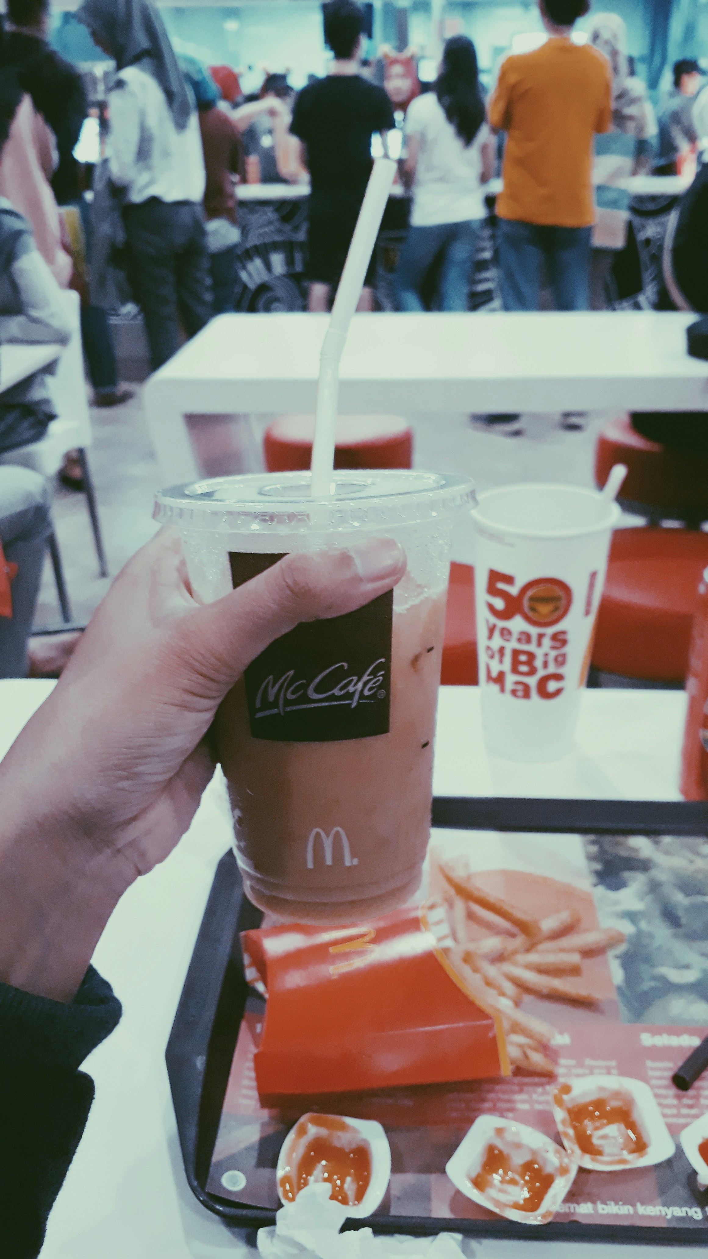 A person holding a McCafé iced coffee drink with a straw in front of a crowded fast-food restaurant. The table holds a tray with McDonald's french fries and sauce containers. Several people are standing in line at the counter.