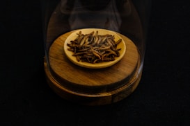 A small wooden bowl containing dried mealworms is placed on a wooden base, all under a transparent glass cover against a dark background.