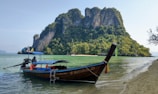 A traditional long-tail boat is moored near the shore of a tropical beach. In the background, a lush, green island with steep cliffs rises dramatically from the water. The water is a clear turquoise, and the sky is bright and clear.