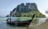 A traditional long-tail boat is moored near the shore of a tropical beach. In the background, a lush, green island with steep cliffs rises dramatically from the water. The water is a clear turquoise, and the sky is bright and clear.