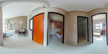 A panoramic view of an interior featuring a modern living room and dining area on the left, separated by a wall with a wooden door leading to a bedroom. The living room has a sofa and a TV unit, while the dining area has a table with chairs. There are two additional rooms visible, one appearing to be a bathroom with a tiled shower section, and another unfinished room with a bed. Natural light enters through windows.