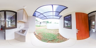 A modern, small kitchen opens to a partially covered outdoor area. The kitchen features a sleek stainless steel sink and countertops with light-colored cabinetry above. Large windows allow natural light to fill the space. The outdoor area is covered with a translucent roof and bordered by a white wall. A wooden door with a metallic handle separates the kitchen from another room.