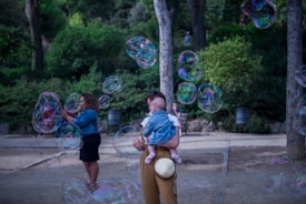 A serene outdoor scene in a park with large, colorful soap bubbles floating through the air. A person holding a baby, both dressed in casual attire, stands in the foreground. In the background, another person is playfully engaged with the bubbles. The park is lush with green trees and shrubs, creating a peaceful, natural environment.