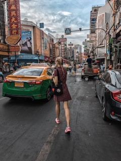 A city street bustling with activity, featuring numerous colorful billboards displaying various advertisements. Traffic consists of cars and taxis navigating the street, while a woman in a checkered dress walks on the road's edge. The atmosphere is lively, with buildings on both sides exhibiting vibrant signage.