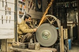 A man is seated while sharpening a tool on a large, spinning grindstone in a workshop. Sparks are flying from the tool being sharpened. The space is filled with various knives and tools displayed on the walls, along with other equipment and workshop items.