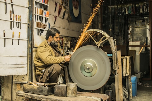 A man is seated while sharpening a tool on a large, spinning grindstone in a workshop. Sparks are flying from the tool being sharpened. The space is filled with various knives and tools displayed on the walls, along with other equipment and workshop items.