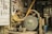 A man is seated while sharpening a tool on a large, spinning grindstone in a workshop. Sparks are flying from the tool being sharpened. The space is filled with various knives and tools displayed on the walls, along with other equipment and workshop items.