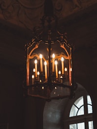 An ornate, vintage chandelier with multiple candle-shaped light bulbs hangs in a dimly lit room. The elaborate metalwork features intricate designs, and soft yellow light casts a warm glow, highlighting the rich textures of the surrounding architecture.