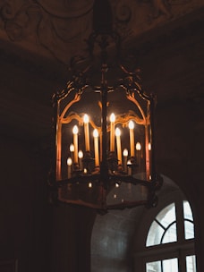 An ornate, vintage chandelier with multiple candle-shaped light bulbs hangs in a dimly lit room. The elaborate metalwork features intricate designs, and soft yellow light casts a warm glow, highlighting the rich textures of the surrounding architecture.